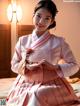 A woman in a pink and white hanbok sitting on a bed.